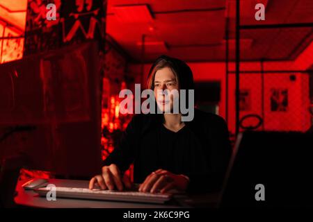 Front view of focused hacker man in black sweatshirt using laptop and desktop computer to breaking network system, from dark basement room with blue Stock Photo