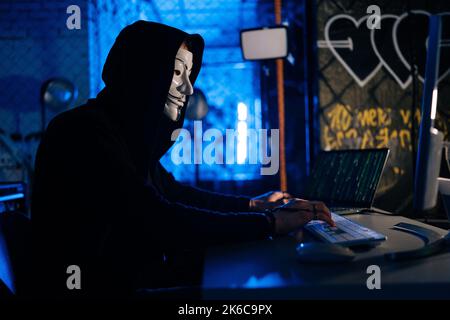 Dangerous hacker male wearing anonymous mask breaking computer system and typing password for hacking bank account using laptop and desktop pc. Stock Photo