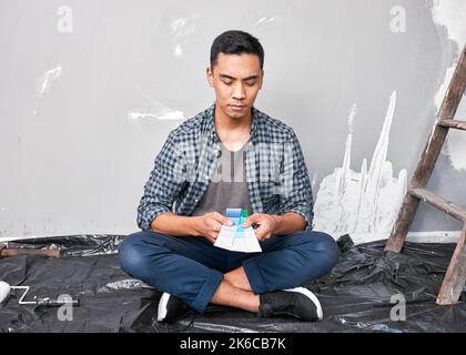 A young Asian man sits on the floor choosing paint colour from swatches home DIY Stock Photo