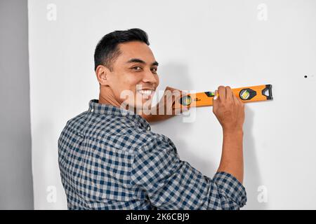 An Asian man smiles while using spirit level against wall doing home DIY Stock Photo