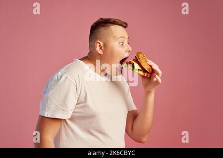 Hungry overweight teen boy in white t-shirt biting delicious hamburger isolated on pink background. Fast food, taste, body positive, emotions Stock Photo