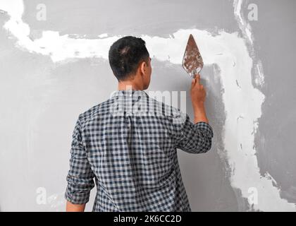 An Asian man smooths plaster over cracks in wall with a trowel home fix DIY Stock Photo