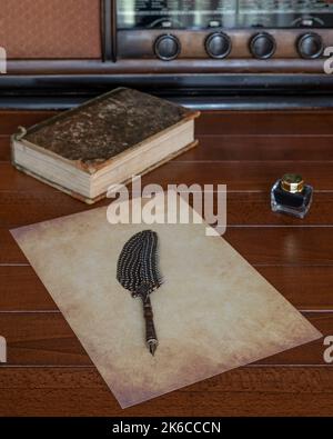 A sheet of antiqued paper with a fountain pen with a feather on it, on a wooden table with a bottle of ink next to it and an ancient radio and an old Stock Photo