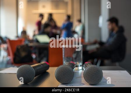 Three microphones over a gray table and on the background a group of people talking and discussing at a conference. Stock Photo