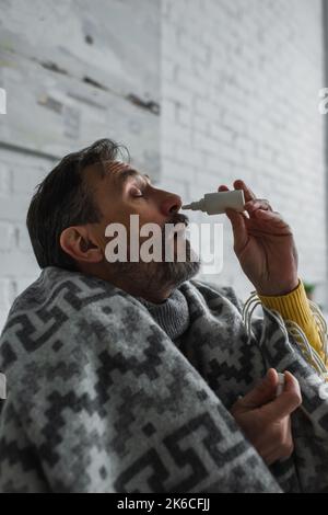 ill man suffering from runny nose and using nasal spray while sitting under warm blanket,stock image Stock Photo