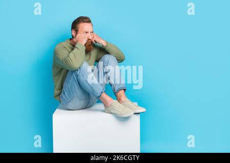 Full size photo of handsome young guy sit offended annoyed depressed white platform wear khaki outfit isolated on cyan color background Stock Photo
