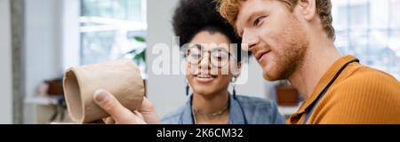 bearded and redhead man checking clay cup near happy african american girlfriend in eyeglasses, banner,stock image Stock Photo