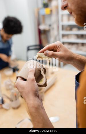 Cropped view of man holding clay cup and sponge during date with blurred african american girlfriend in pottery workshop,stock image Stock Photo