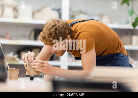 Redhead master in apron creating clay cup in pottery workshop,stock image Stock Photo
