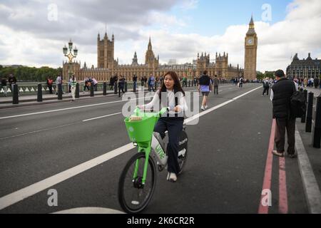 A young Asian women rides a Lime electric bike over Westminster Bridge. The bridge is closed to cars during Queen lying in State ceremony. Stock Photo