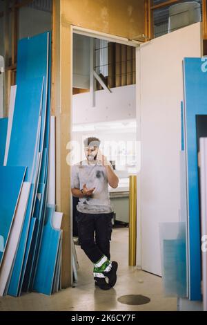 Full length of young male carpenter talking on smart phone while standing at doorway of workshop Stock Photo