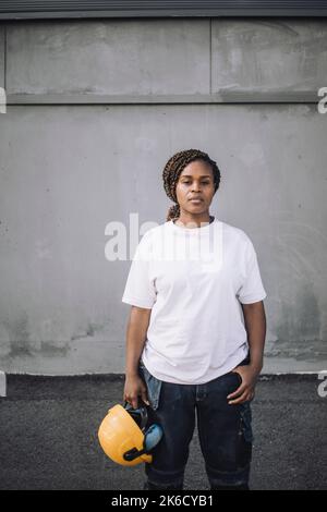 Female construction worker standing with hardhat against wall Stock Photo