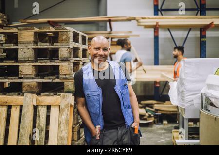 Smiling bald carpenter with hands in pockets leaning on wooden rack in warehouse Stock Photo