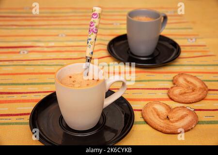 two cups of coffee next to some palm-shaped cookies on yellow tablecloth Stock Photo