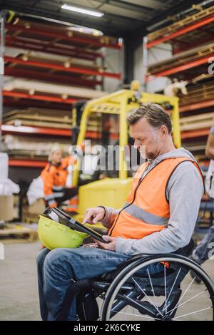 Male carpenter with disability using digital tablet sitting on wheelchair in warehouse Stock Photo