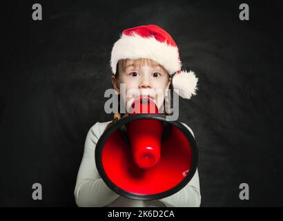 Happy child girl in Santa hat speaking through a megaphone on blackboard background Stock Photo