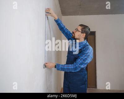 Side view of serious man using measuring tape in room with underfloor heating pipes. Designer in shirt taking measurements while working on renovation of apartment. Stock Photo