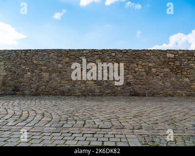 Old stone wall in front of a blue sky with clouds. Empty cobblestone street and an old masonry. Medieval building exterior in a rural area. Stock Photo
