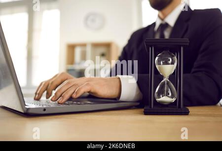 Man trying to meet deadline and working on laptop computer at desk with hour glass Stock Photo