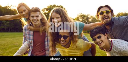 Group of cheerful young friends having fun with piggyback outdoors in park on summer day. Stock Photo