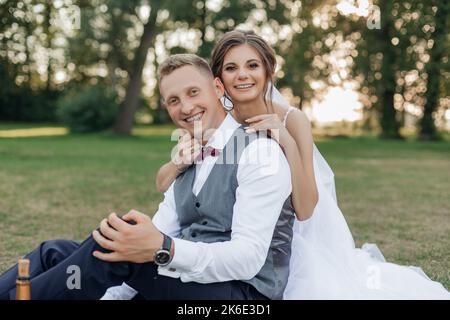 Portrait of beautiful wedding couple sitting on green grass in park in summer. Young cheerful woman bride hugging groom. Stock Photo