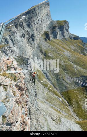 Climber Ascends The 540° Grade Ladder In The Gemmiwand Via Ferrata. In The Background Plattenhörner, Blue Sky. Leukerbad, Valais, Switzerland. Stock Photo