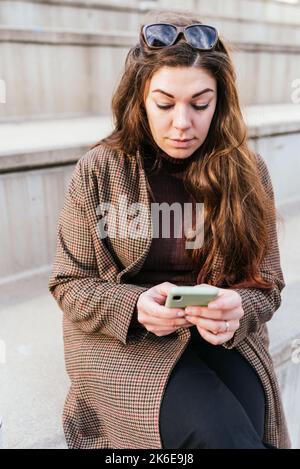 Female in outerwear with long hair browsing smartphone while sitting on steps on city street at night Stock Photo