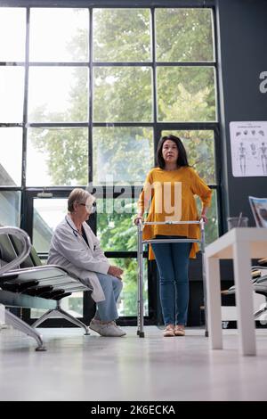 Elderly doctor attending physiotherapy with asian patient, doing rehabilitation exam and trying to walk with walking frame. Injured woman with leg fracture working on physical recovery. Stock Photo