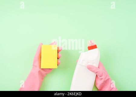 Cleaner's hand in pink rubber protective glove holding bottle with detergent and yellow sponge on green background. Top view. Stock Photo