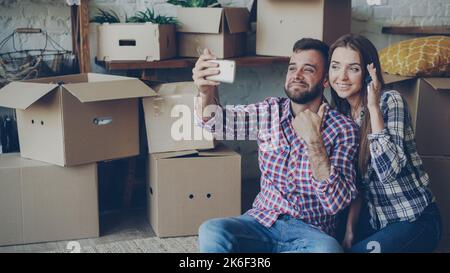Happy young couple is making video call with smartphone after relocation. They are greeting friends, showing new house keys and many boxes, chatting and smiling. Stock Photo