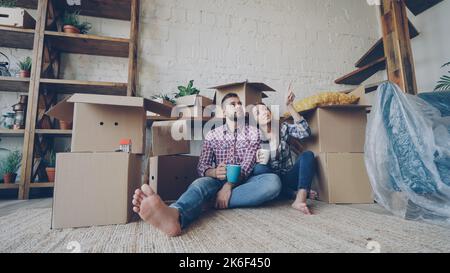 Adorable young couple is talking and holding mugs while sitting on floor of new flat after relocation. Numerous boxes, packed furniture and plants are visible. Stock Photo