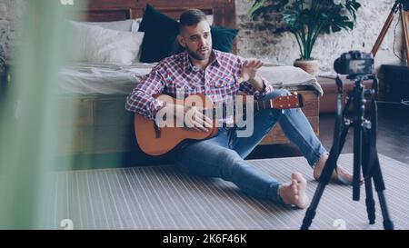 Handsome man blogger and guitarist is recording video lesson about guitar in bedroom using camera. Bearded guy is greeting his followers, talking and showing guitar parts. Stock Photo