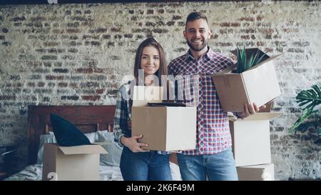 Portrait of happy young couple standing in bedroom in new house, holding carton boxes, smiling and looking at camera. Family, relationship and relocation concept. Stock Photo