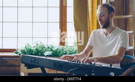 Cheerful young man amateur musician in casual clothes is playing keyboard at home. Musical instruments, attractive people, hobby and creativity concept. Stock Photo