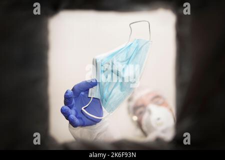Hand in medical gloves throwing out discarded, used protective masks in the trash bin after quarantine. A medical technician throws away a face mask. Stock Photo