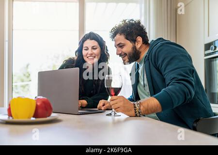 Happy couple of young newlywed using laptop in the kitchen surfing internet content and drinking red wine against a bright window - people and technol Stock Photo