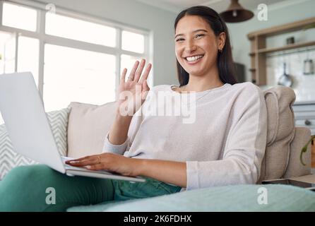 Shes the face behind the blog. a woman waving while using her laptop on the couch at home. Stock Photo