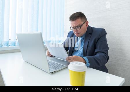 A male employee in a blue jacket works with documents and on a computer in a bright office. Surprised emotion on the face. Stock Photo