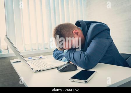 Tired businessman sleeping in his office. A man in a blue jacket sleeps in the morning at the workplace. The employee fell asleep at the table in fron Stock Photo