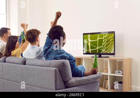 Happy diverse friends sitting on sofa, watching football on TV and drinking beer Stock Photo