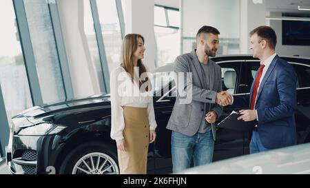 Young bearded man is buying car for his attractive wife shaking hands with manager and getting key Stock Photo