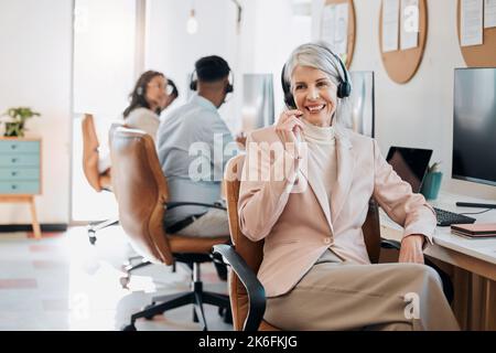 How can we help you today. a mature businesswoman sitting in the office and wearing a headset while her colleagues work behind her. Stock Photo