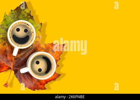 Coffee cups with smiley faces placed on a large pigmented maple leaf isolated on yellow with copy space. Cosy autumn background. Stock Photo