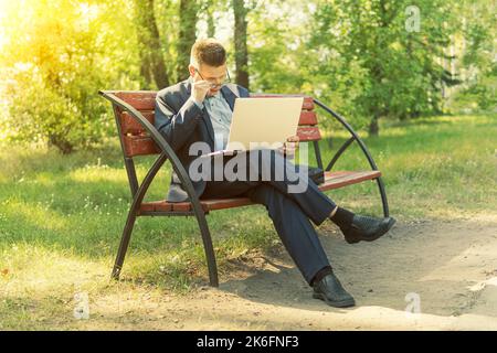Freelancer programmer sitting at laptop on a bench in Park outdoors. businessman working on his laptop connect to wireless internet in rural area. Man Stock Photo
