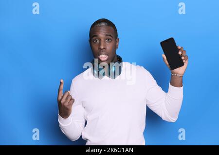 Emotional black man holds a smartphone in his hands, showing his finger up in a blank space. Blue background Stock Photo
