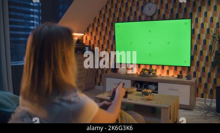 Woman switching channels with remote control. Couple sitting on sofa in living room, talking, watching breaking news, movie or series on TV. Resting at home at night. Green screen. Chromakey. Stock Photo
