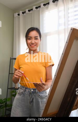 Image of female painter holding paintbrush and palette standing in front of canvas and smiling to camera Stock Photo