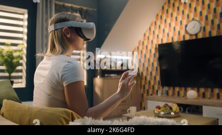 Woman wearing VR headset, sitting on sofa in cozy living room in midday, gesturing, playing video game in virtual reality, testing VR goggles, spending weekend at home. Cyberspace and metaverse. Stock Photo