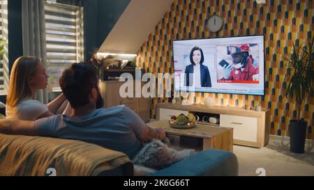Couple sitting on sofa in living room together, talking, watching TV news about disaster, terrorist atack, collapsed building and rescuing people from rubble. Spending time at home. TV live broadcating. Stock Photo