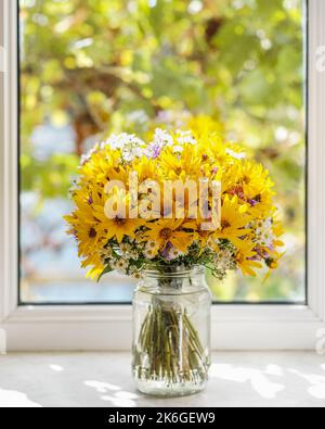 Bouquet of yellow, violet and white wild flowers in a jar against window. Autumn composition. Stock Photo
